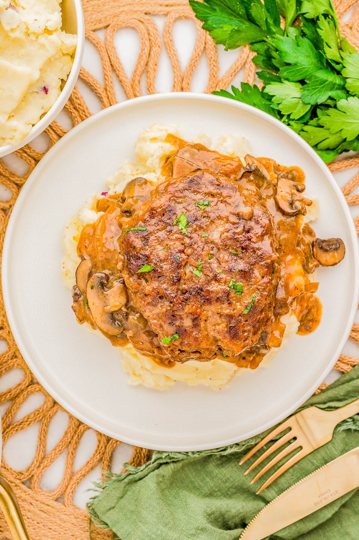 A hamburger steak with mushroom gravy on mashed potatoes, garnished with parsley, is served on a white plate. Accompanied by a fork, knife, a green napkin, and a bowl of mashed potatoes.