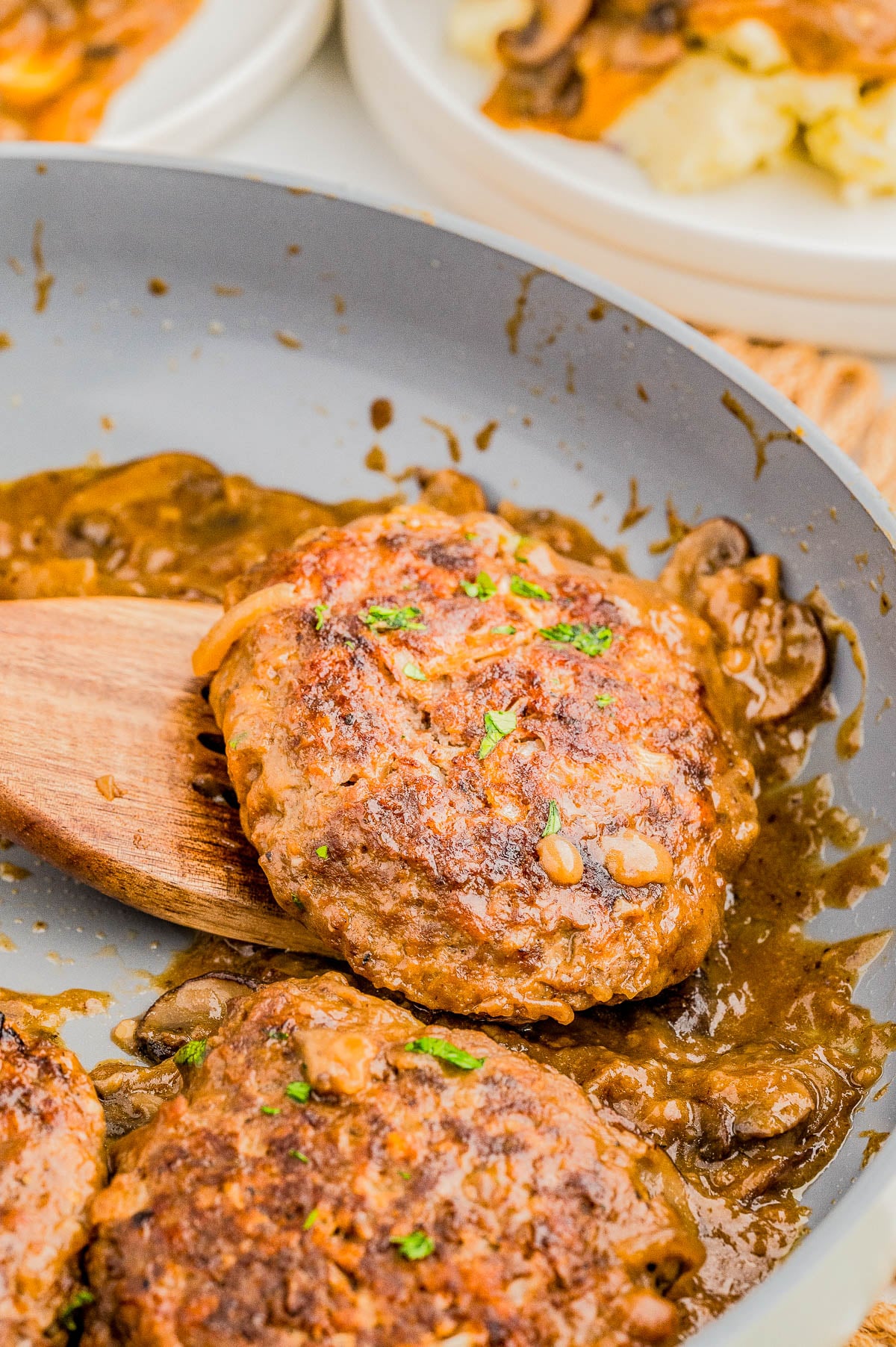 Salisbury steak patties in a pan with brown mushroom gravy, garnished with chopped parsley. A wooden spatula is lifting one patty. Plates in the background.