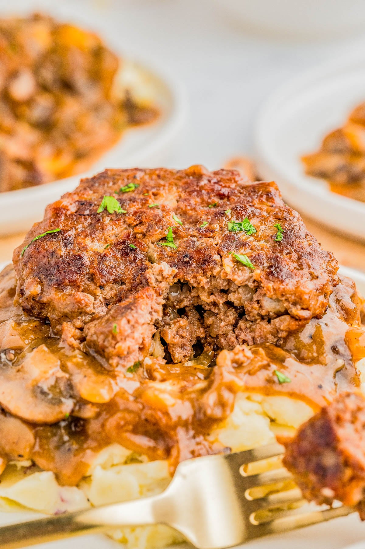 A cooked meat patty topped with gravy sits on mashed potatoes, garnished with herbs. A fork is placed nearby.