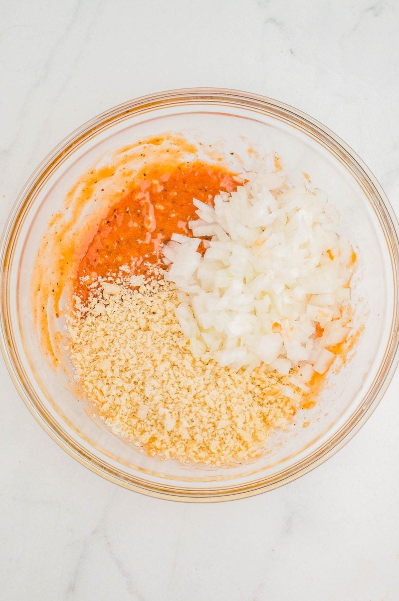 A clear glass bowl containing a mixture of chopped onions, breadcrumbs, and a red liquid on a white marble surface.