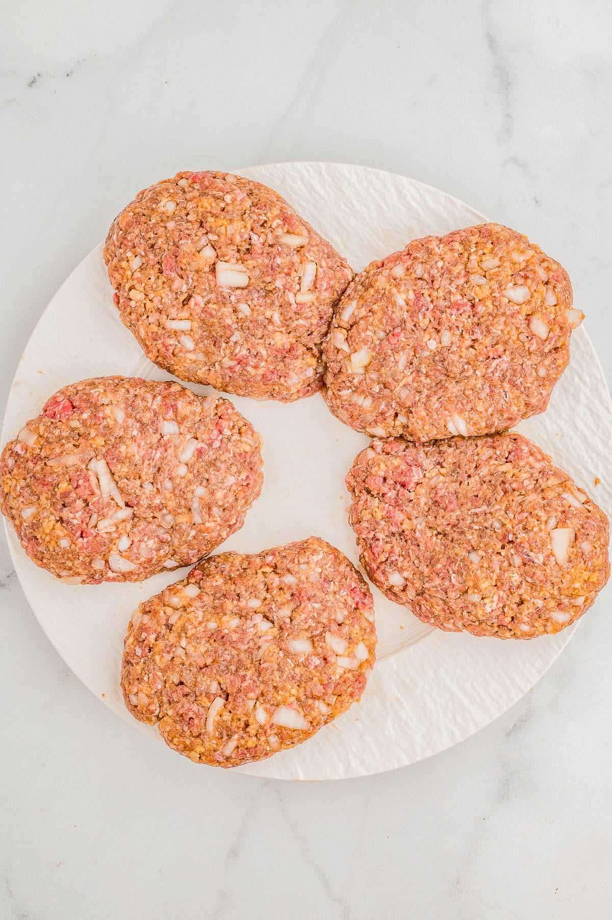 Five raw hamburger patties with visible onion slices on a white plate, placed on a marble surface.