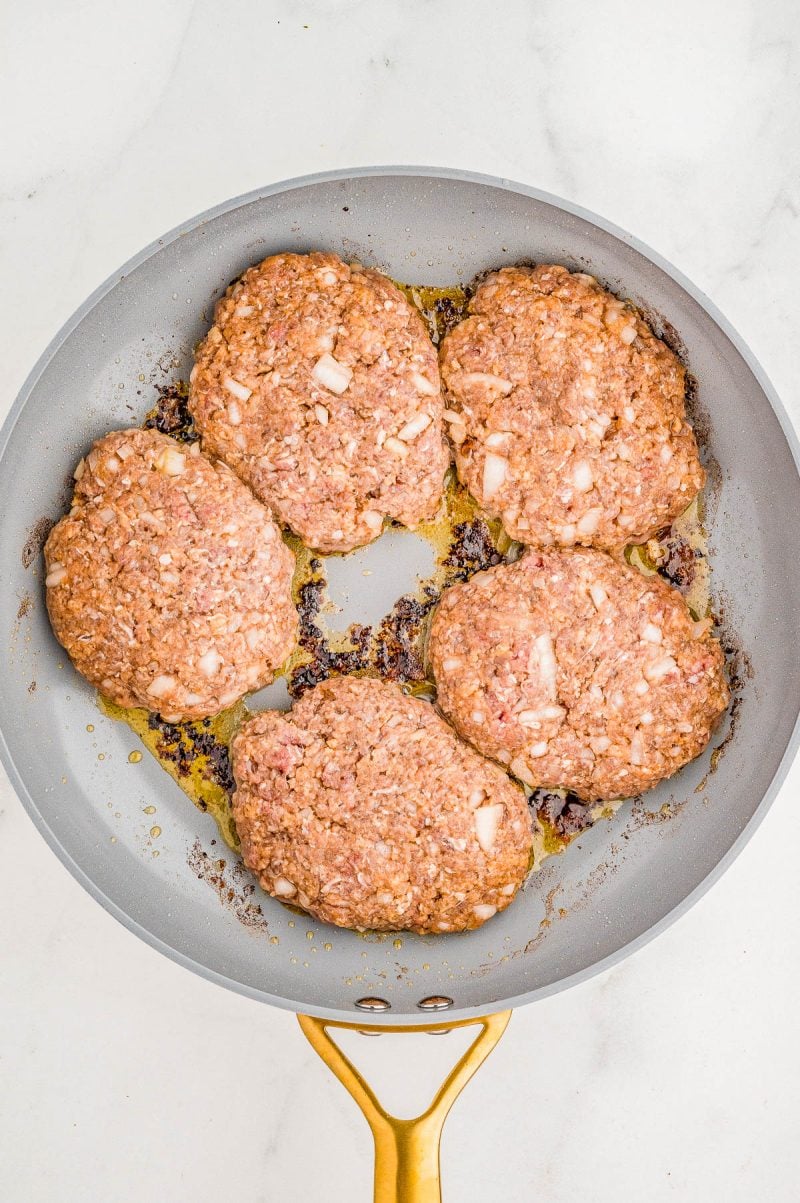 Four raw hamburger patties with diced onions in a frying pan on a stovetop.