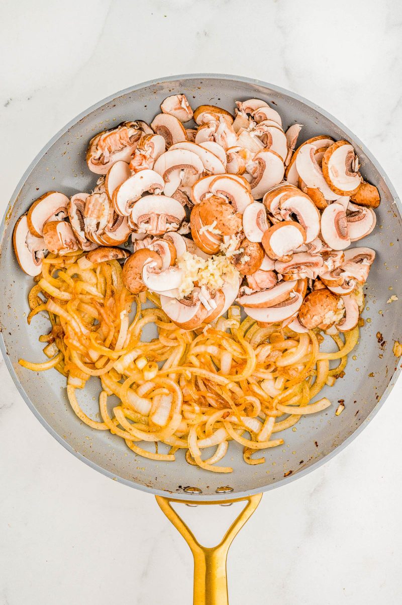 A frying pan contains sliced mushrooms, sautéed onions, and minced garlic on a white background.