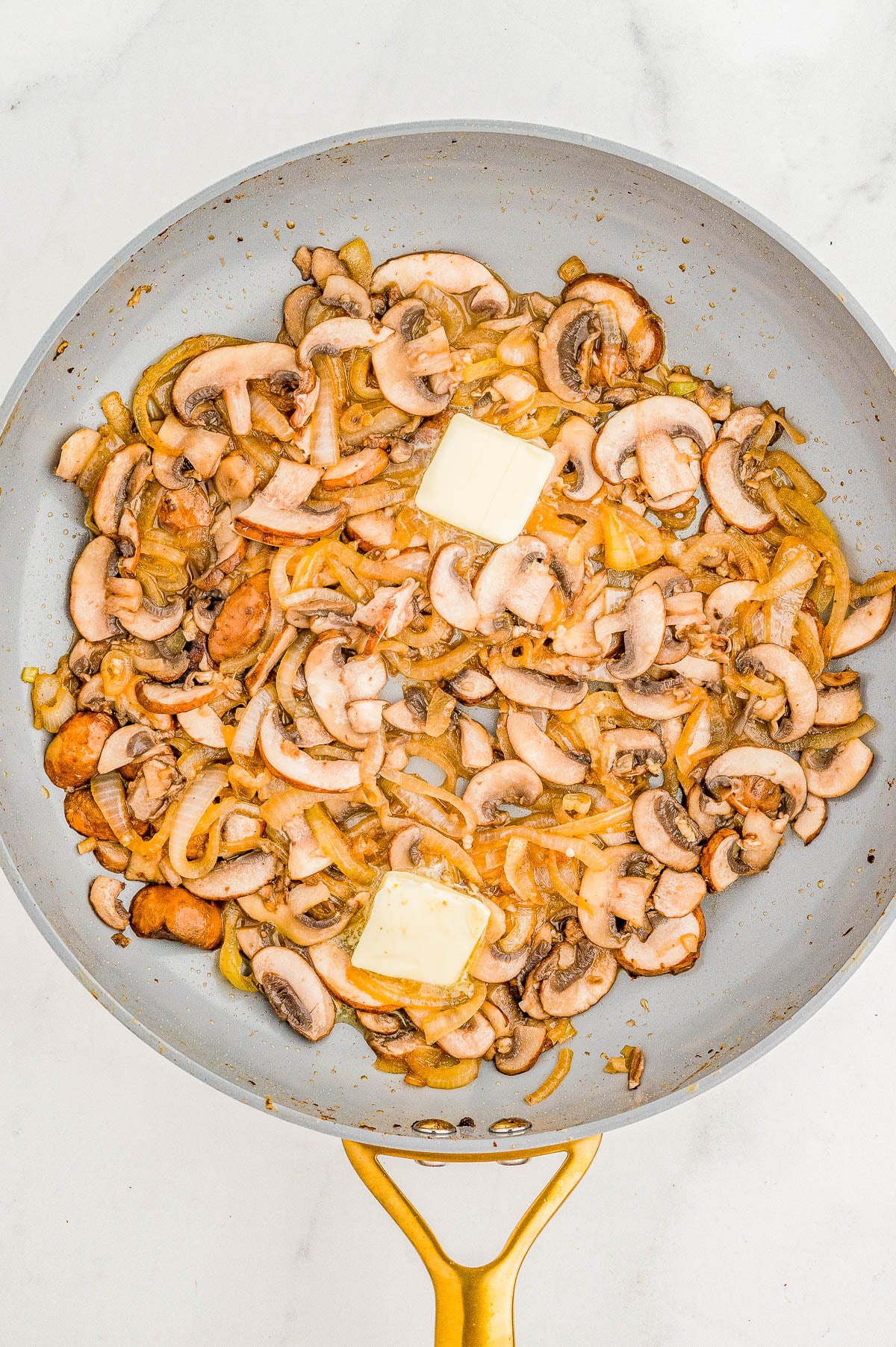 Sautéing sliced mushrooms and onions with two pieces of butter in a frying pan on a stovetop.