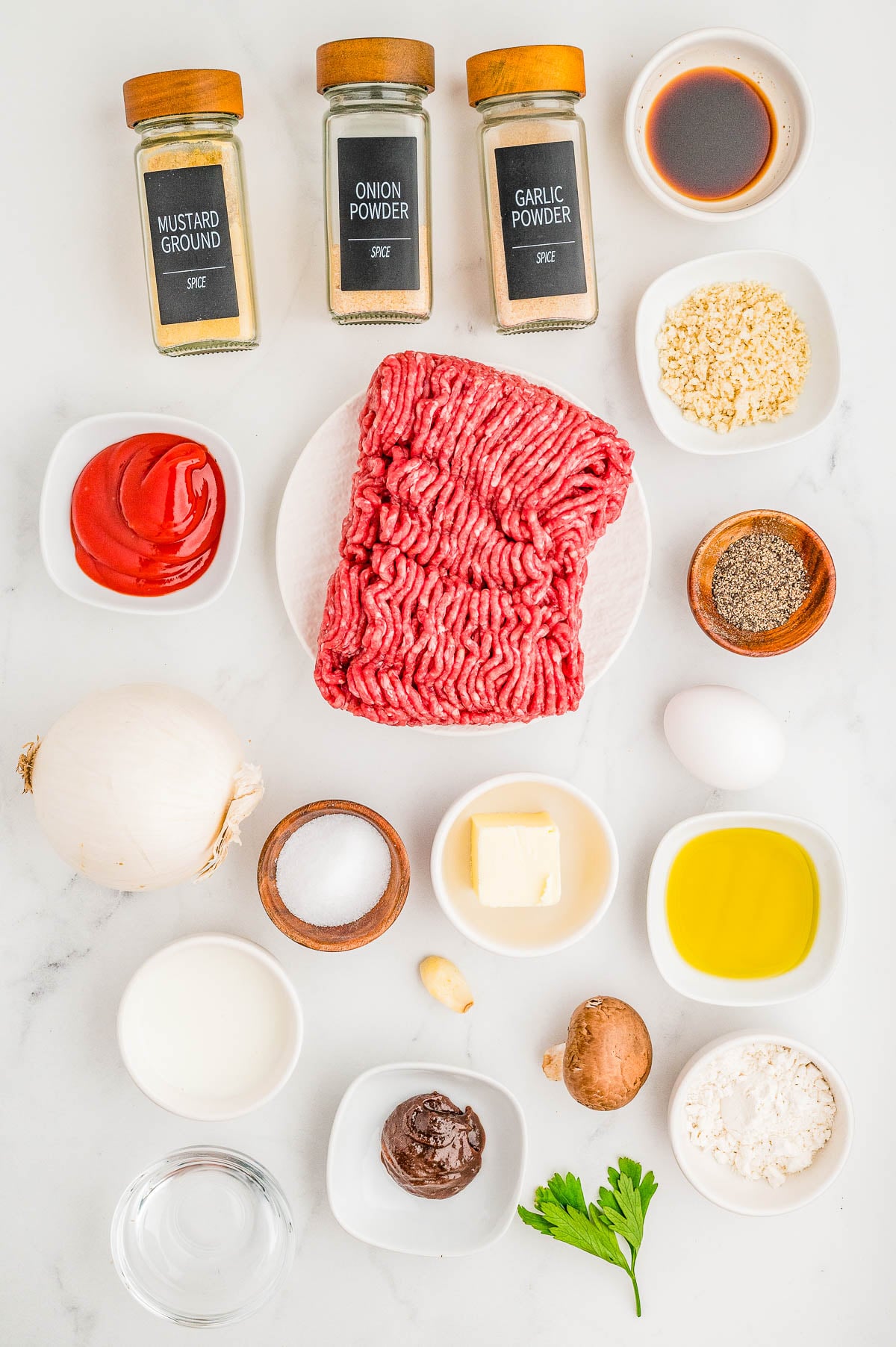 Raw ground meat surrounded by spices, condiments, and ingredients including onion, egg, butter, mushroom, oil, breadcrumbs, and seasonings on a white surface.