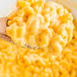 A wooden spoon holds a portion of creamy macaroni and cheese with melted cheese strings, against a background of the same dish.