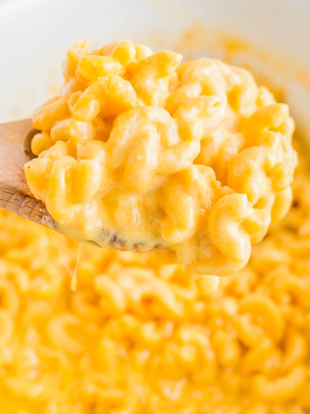 A wooden spoon holds a portion of creamy macaroni and cheese with melted cheese strings, against a background of the same dish.