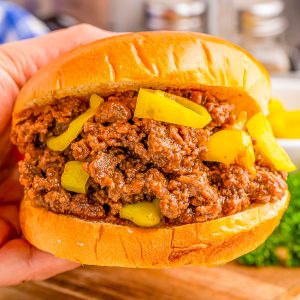 Hand holding a sloppy joe sandwich with beef filling and yellow pepper slices, on a wooden surface.