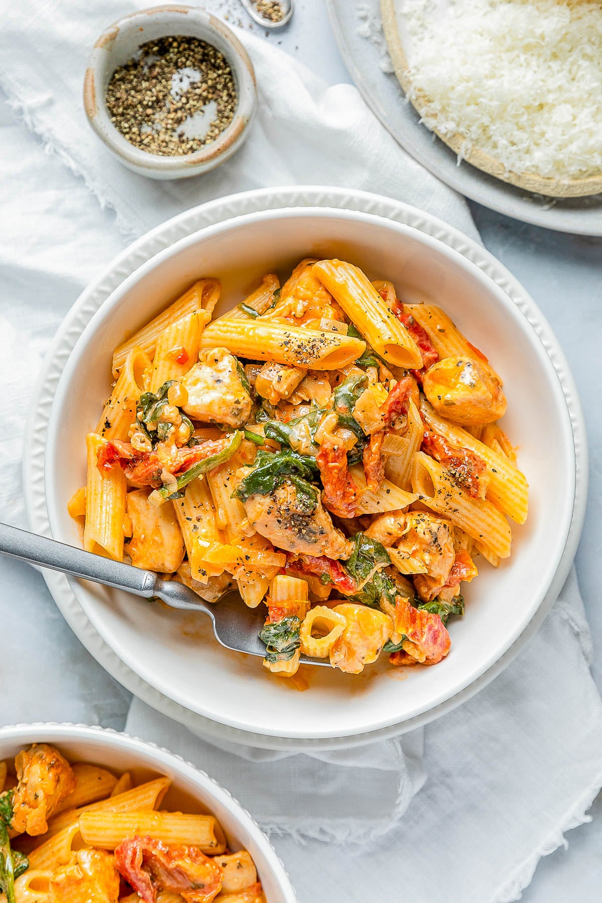 A bowl of creamy penne pasta with chicken, spinach, and tomatoes, garnished with pepper. A fork rests in the bowl. Grated cheese and pepper are in bowls beside it.