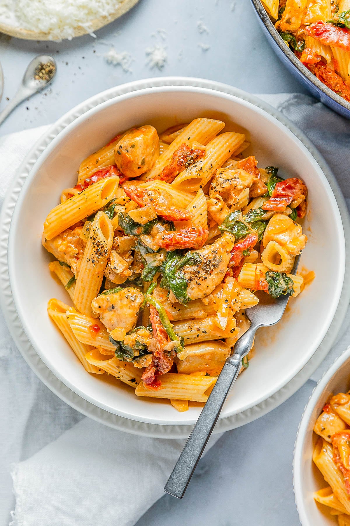 A bowl of penne pasta with creamy sauce, chicken pieces, spinach, and red bell peppers, garnished with black pepper, on a light-colored surface, with a fork placed in the bowl.