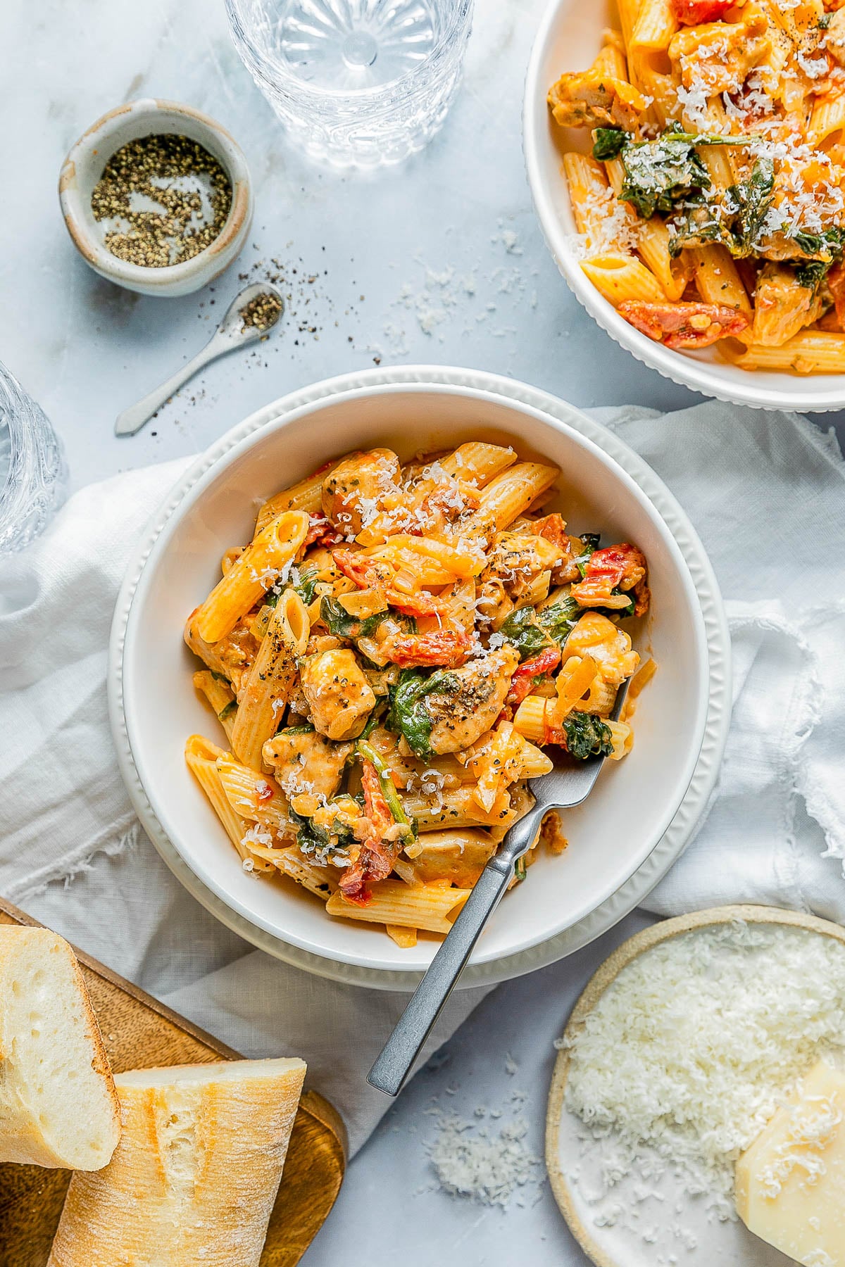 A bowl of pasta with vegetables and grated cheese on top, surrounded by bread, grated cheese, and a bowl of pepper on a white table.