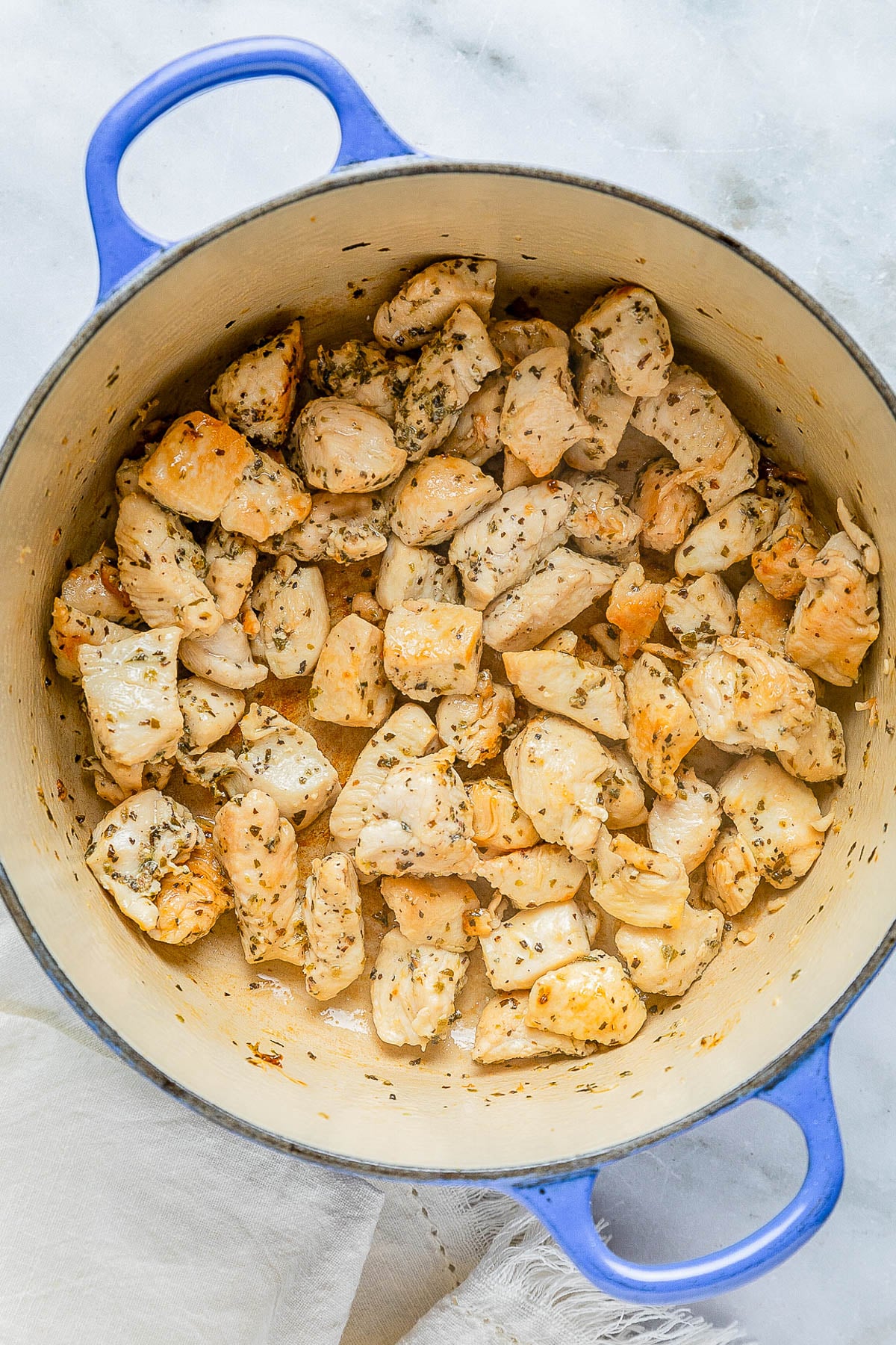 Cooked cubed chicken with herbs in a large round blue pot on a marble surface.