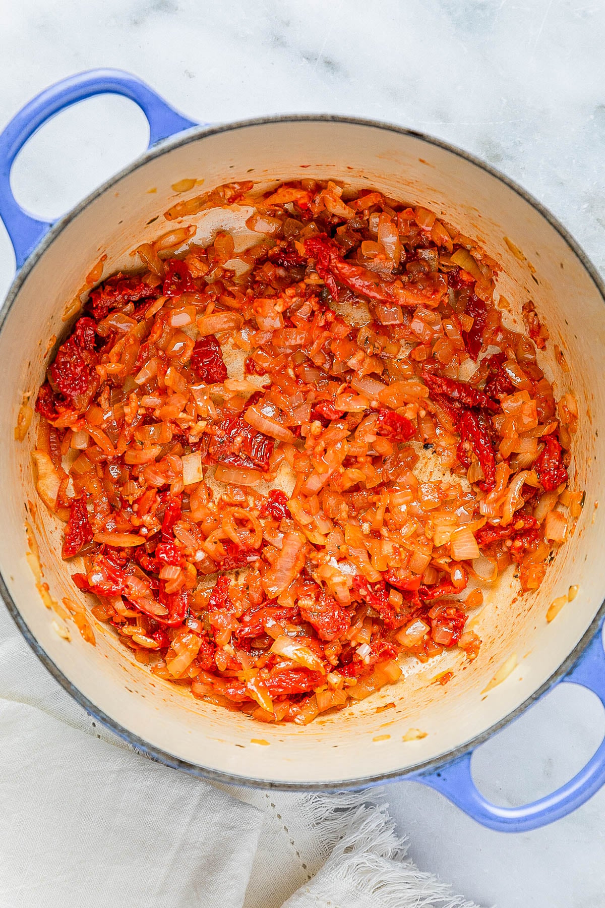 Chopped onions and red peppers are sautรฉed in a large blue pot, sitting on a light countertop.