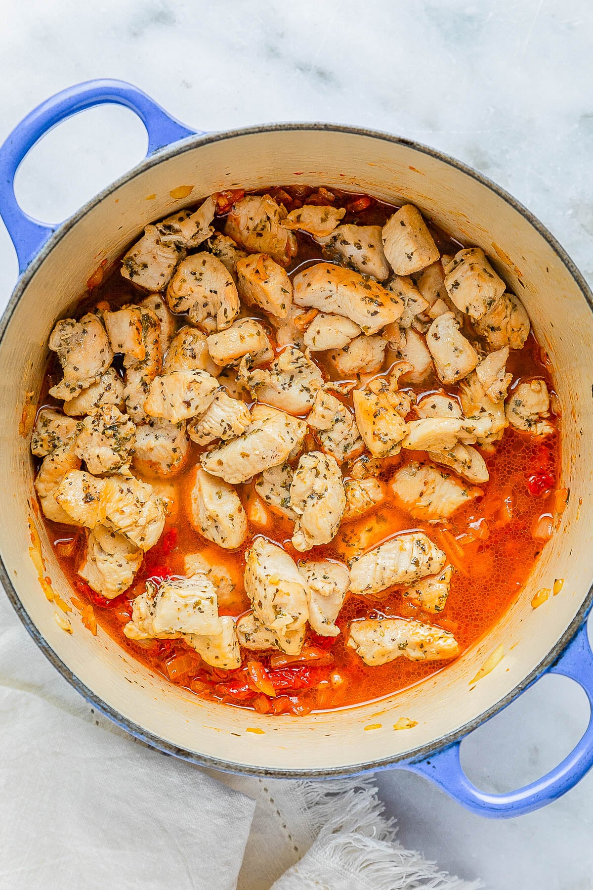 Chunks of cooked chicken with herbs in a blue pot, simmering in a red-orange liquid on a white surface.