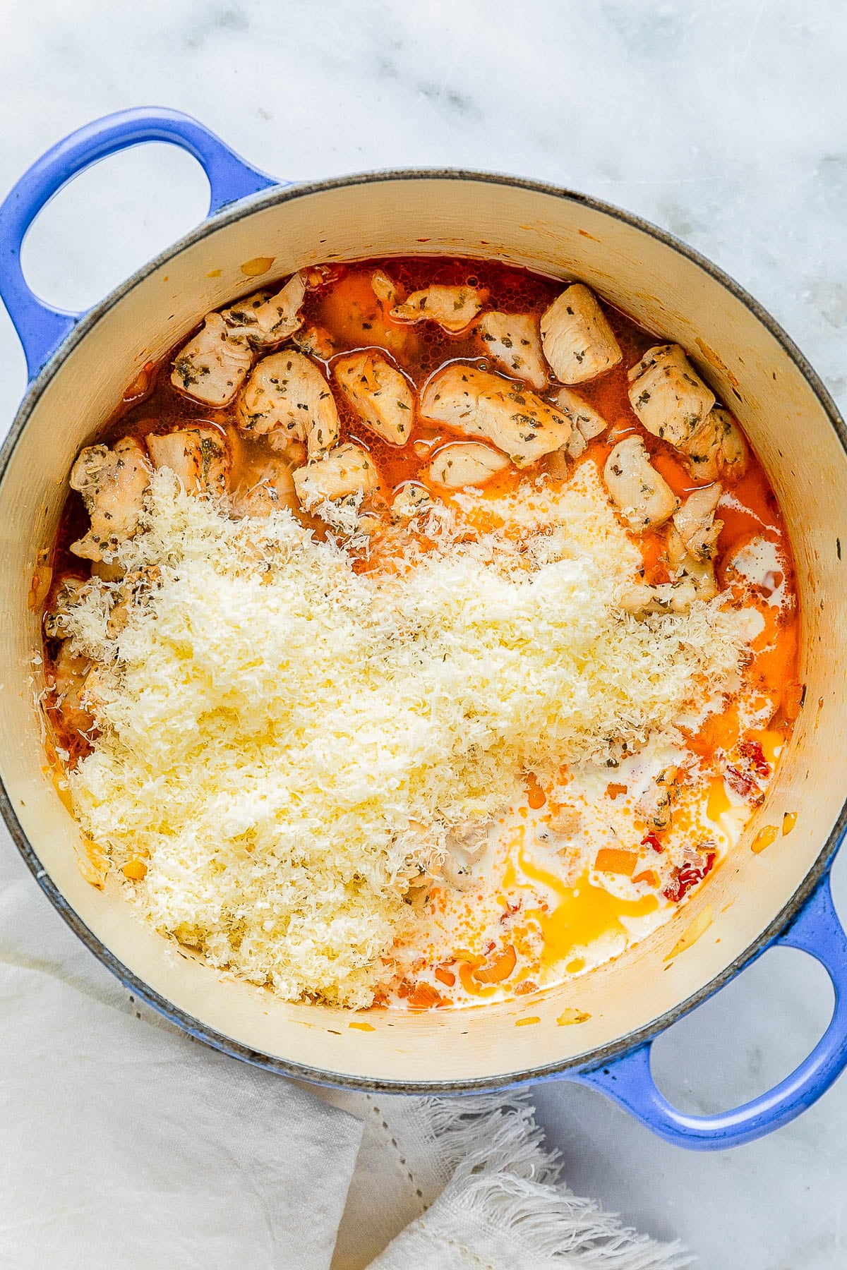 Blue pot with chicken, sauce, and grated cheese, set on a light surface.