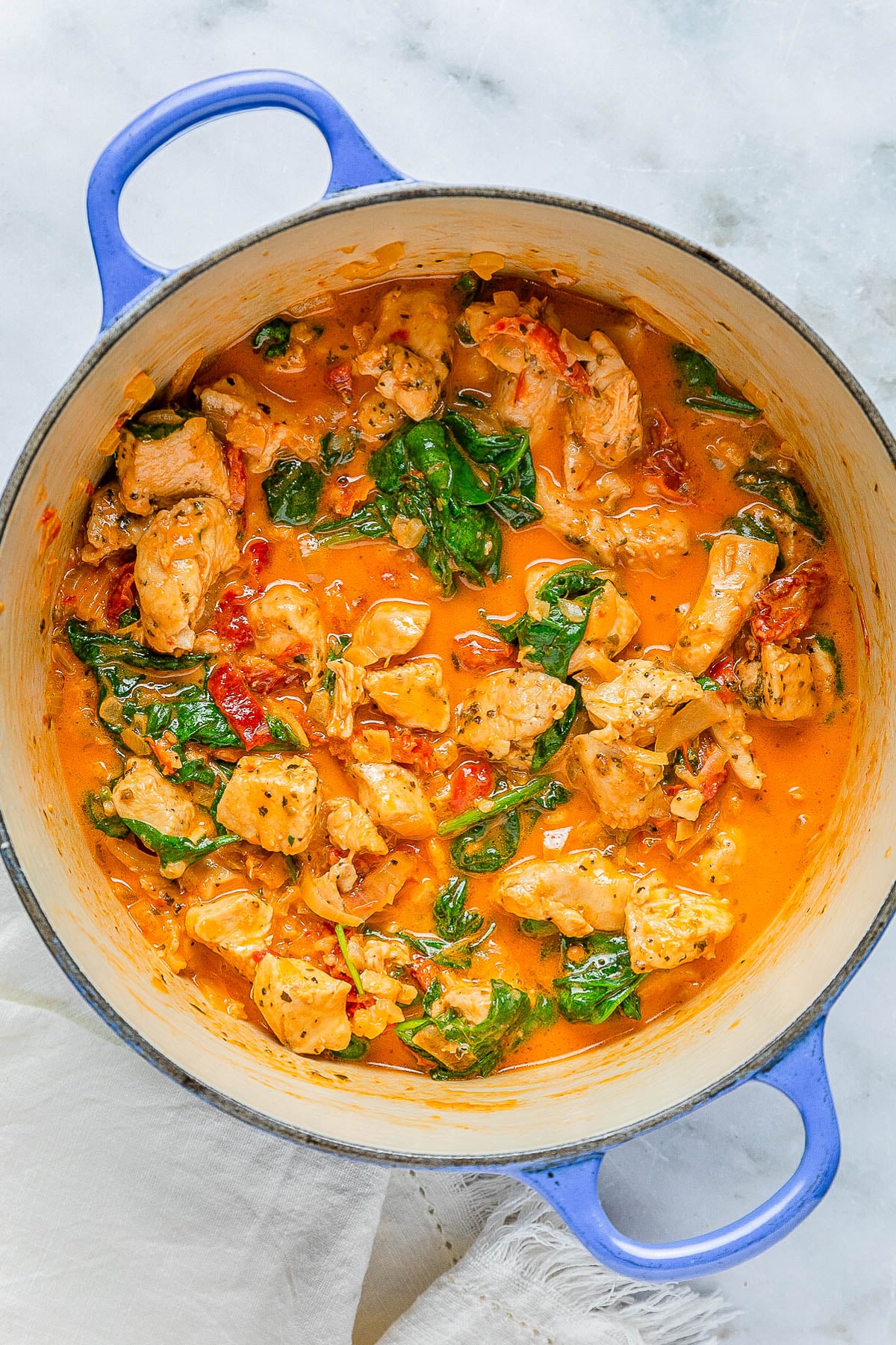 A blue pot with creamy chicken, spinach, and tomatoes in a sauce, placed on a marble surface.