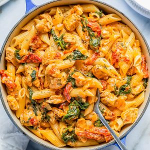 A blue pot filled with creamy pasta featuring penne, chicken pieces, spinach, and cherry tomatoes, with a serving spoon resting inside. Plates and cheese in the background.
