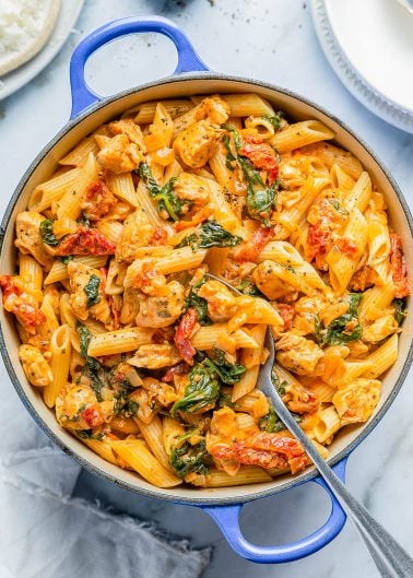 A blue pot filled with creamy pasta featuring penne, chicken pieces, spinach, and cherry tomatoes, with a serving spoon resting inside. Plates and cheese in the background.