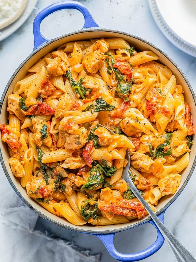 A blue pot filled with creamy pasta featuring penne, chicken pieces, spinach, and cherry tomatoes, with a serving spoon resting inside. Plates and cheese in the background.