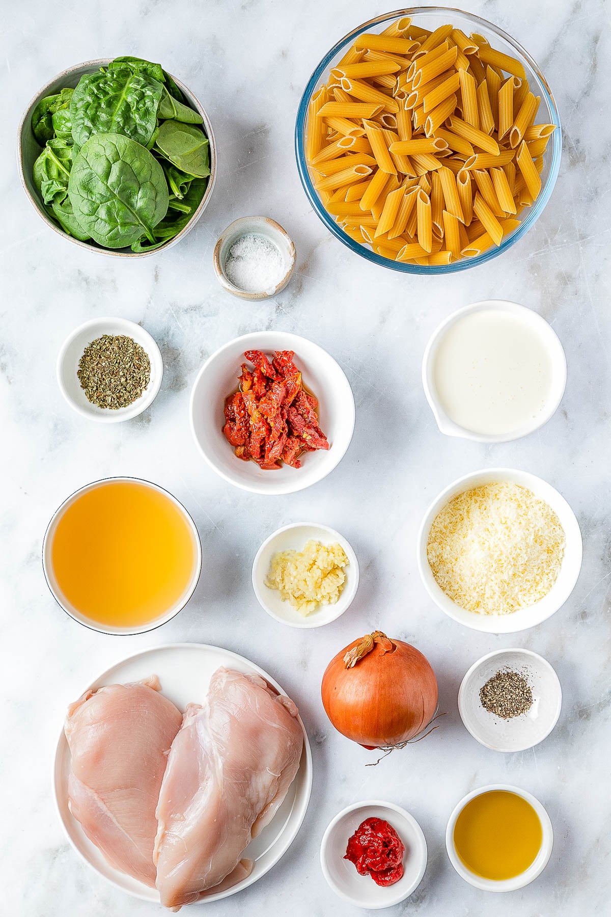 Ingredients for a pasta dish arranged on a surface, including spinach, penne, chicken, sun-dried tomatoes, cream, onion, grated cheese, broth, and various seasonings.