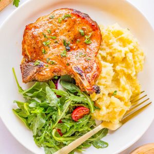 A plate with a grilled pork chop, mashed potatoes, and a green salad with cherry tomatoes.