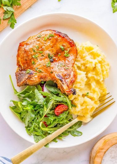 A plate with a grilled pork chop, mashed potatoes, and a green salad with cherry tomatoes.