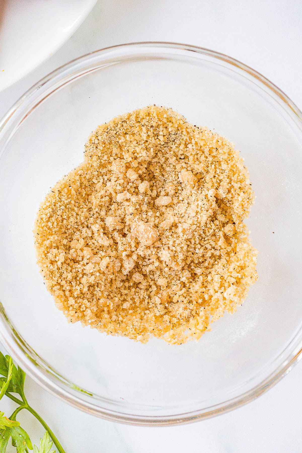 Close-up of light brown breadcrumbs in a transparent glass bowl on a white surface.