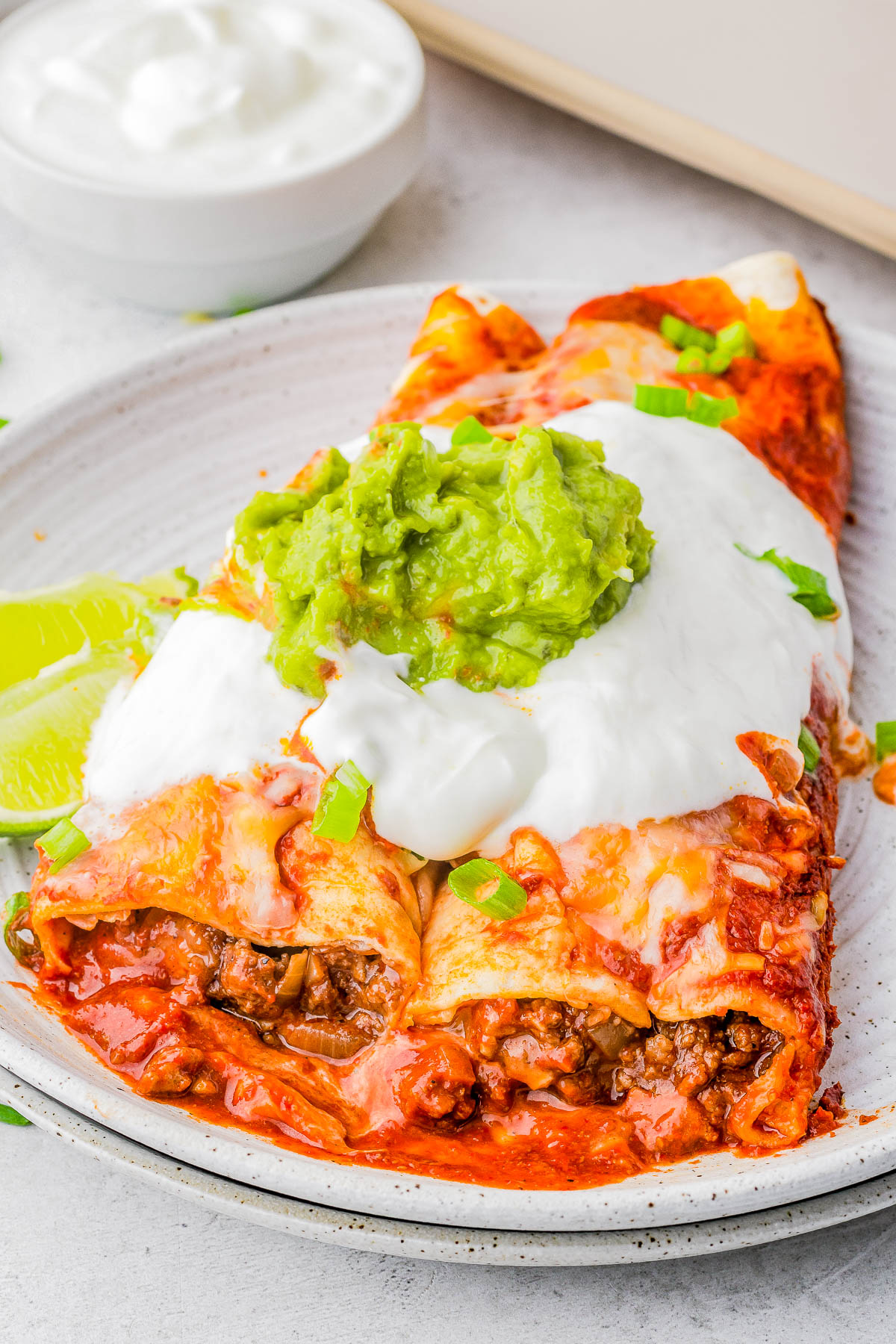 Plate of stuffed enchiladas with red sauce, topped with sour cream and guacamole, garnished with chopped green onions, alongside lime wedges.