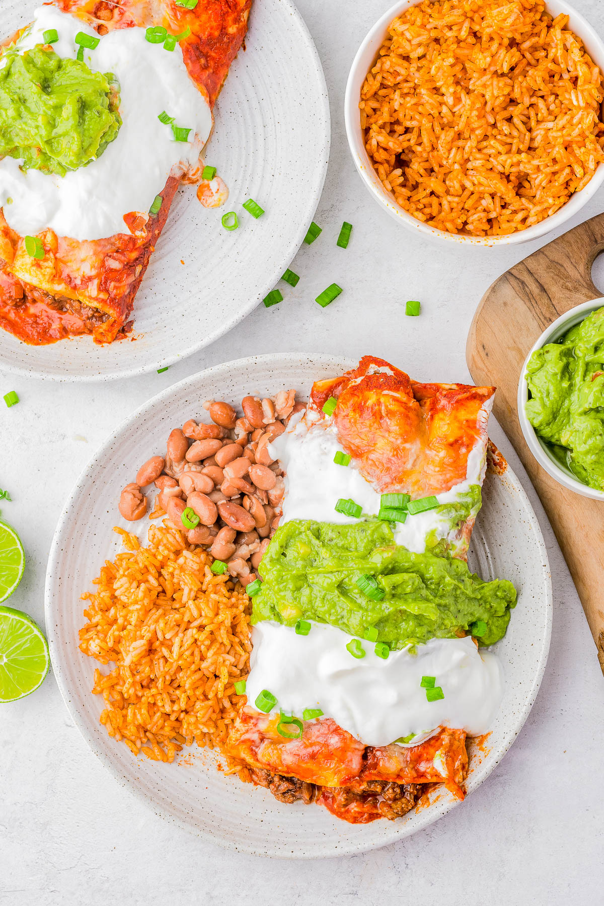Plates of enchiladas topped with sour cream and guacamole, served with Mexican rice and beans. Bowls of additional rice and guacamole are on the side. Lime wedges are present.