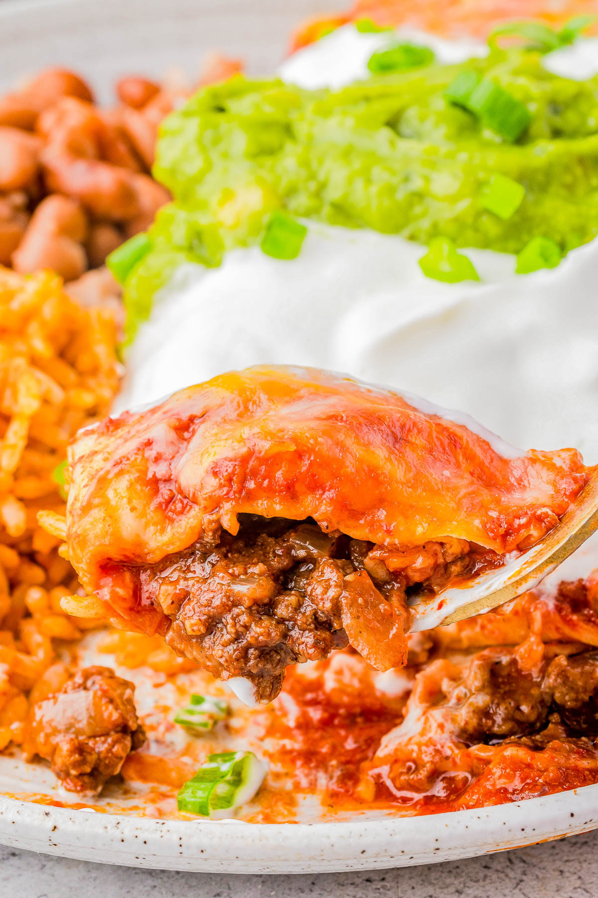 A close-up of a plate with enchilada topped with melted cheese. Rice and beans are in the background, along with sour cream and guacamole garnished with green onions.