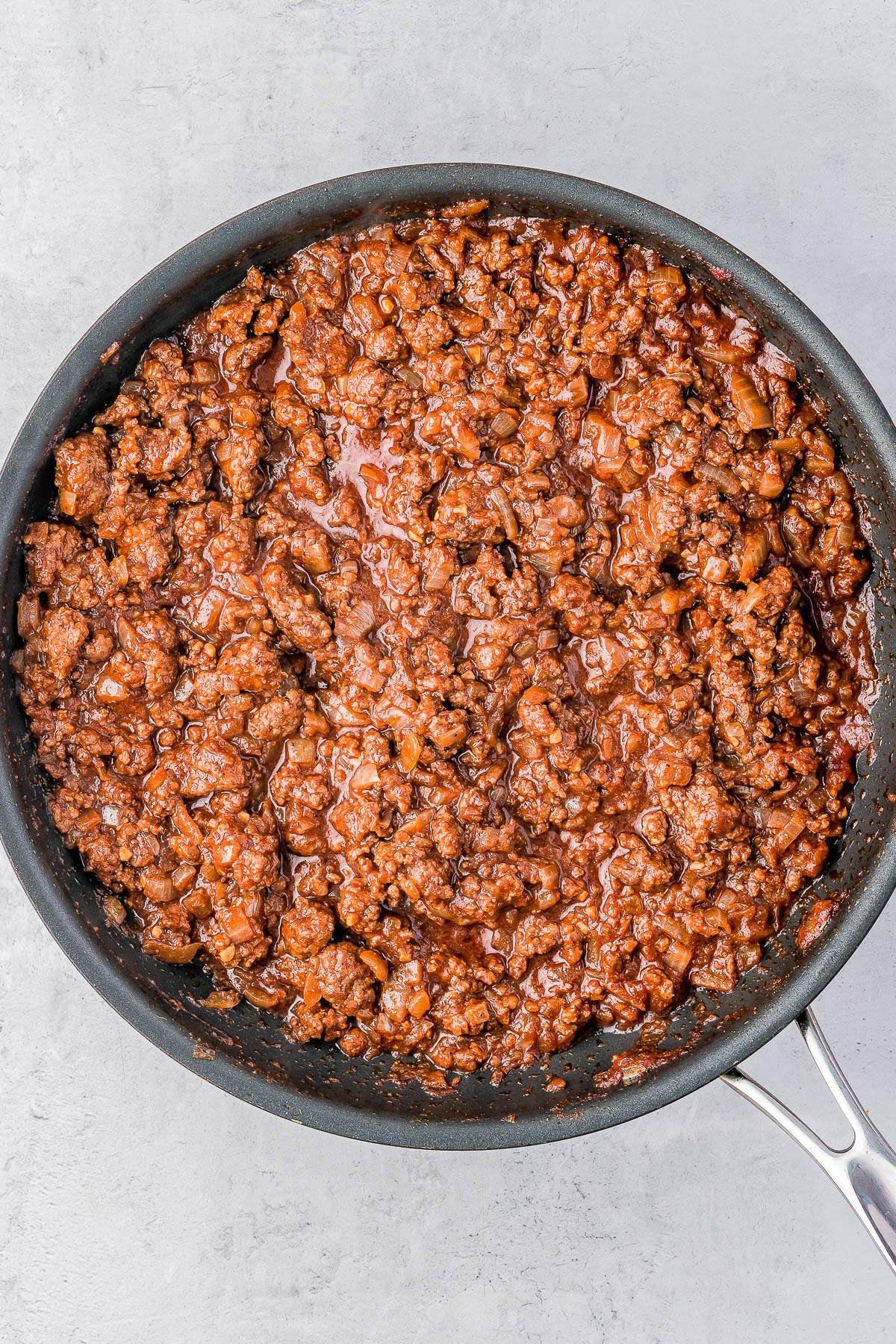 A pan filled with cooked ground beef seasoned with a reddish sauce, resting on a light gray surface.