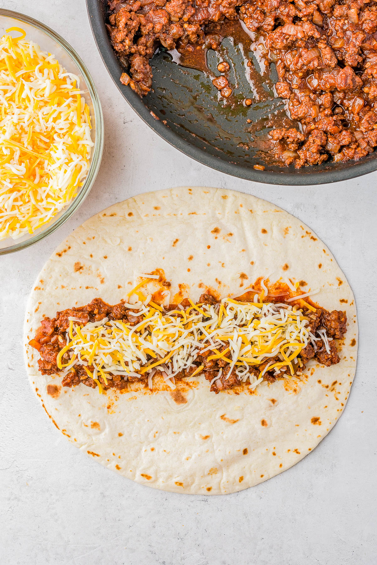 Tortilla on a flat surface with ground meat and shredded cheese. A pan of meat and a bowl of cheese are nearby.
