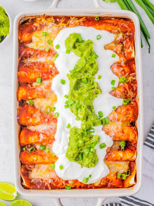 A dish of enchiladas topped with sour cream, guacamole, and chopped green onions. Lime wedges and a bowl of sauce are on the side.