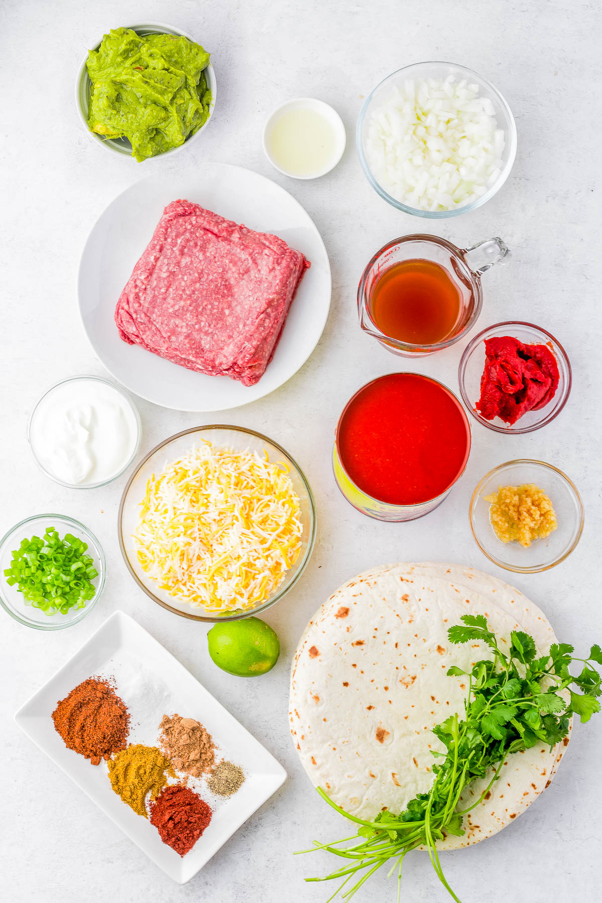 Ingredients for tacos arranged on a table, including ground meat, cheese, tortillas, diced onions, sour cream, avocado, spices, lime, coriander, garlic, vinegar, tomato sauce, and beef stock.