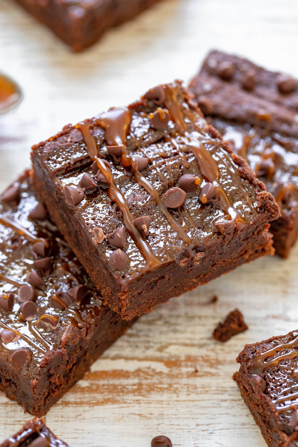 Close-up of stacked chocolate brownies with drizzled caramel and chocolate chips on a light wooden surface.