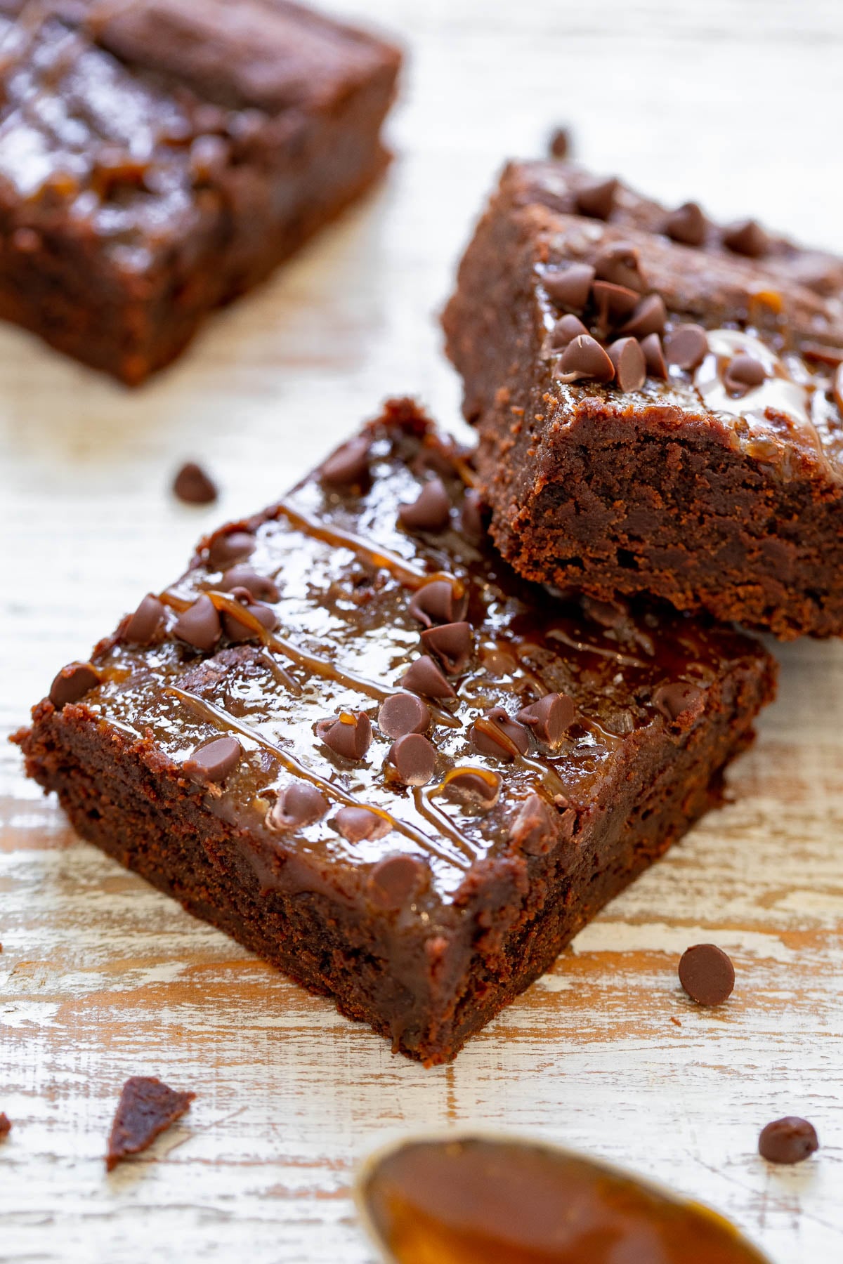 Two chocolate brownies topped with chocolate chips and drizzle, placed on a white wooden surface. A spoon with chocolate sauce is visible in the foreground.