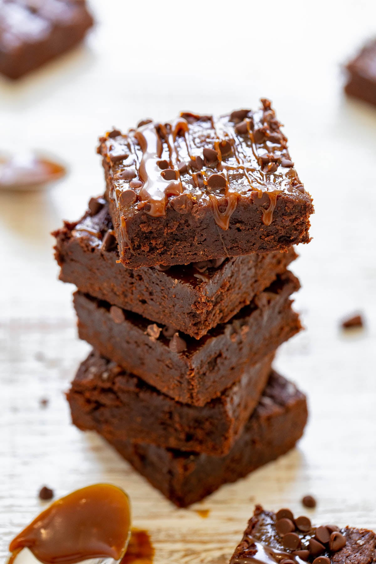 A stack of four chocolate brownies drizzled with caramel sauce and topped with chocolate chips, placed on a light wooden surface.