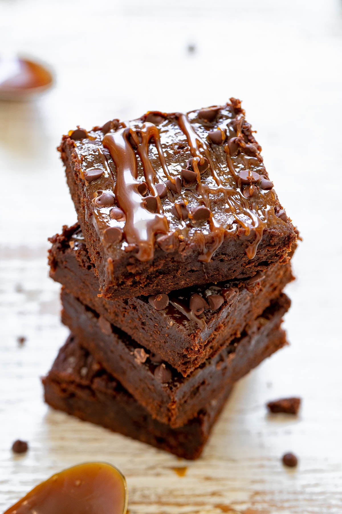 A stack of chocolate brownies with chocolate drizzle and small chocolate chips on top, placed on a white wooden surface.