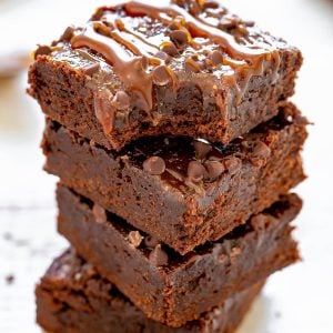 A stack of four chocolate brownies topped with caramel drizzle and chocolate chips on a wooden surface.