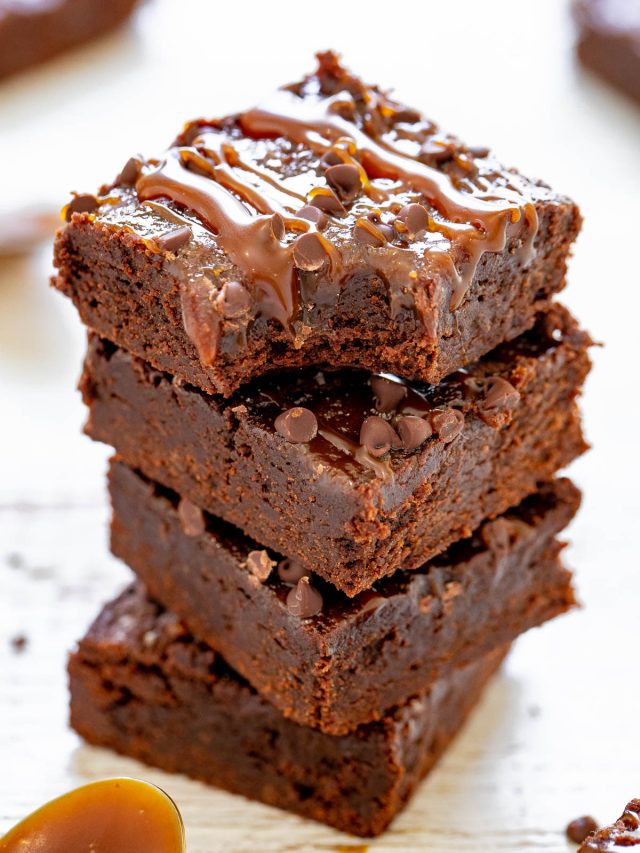 A stack of four chocolate brownies topped with caramel drizzle and chocolate chips on a wooden surface.