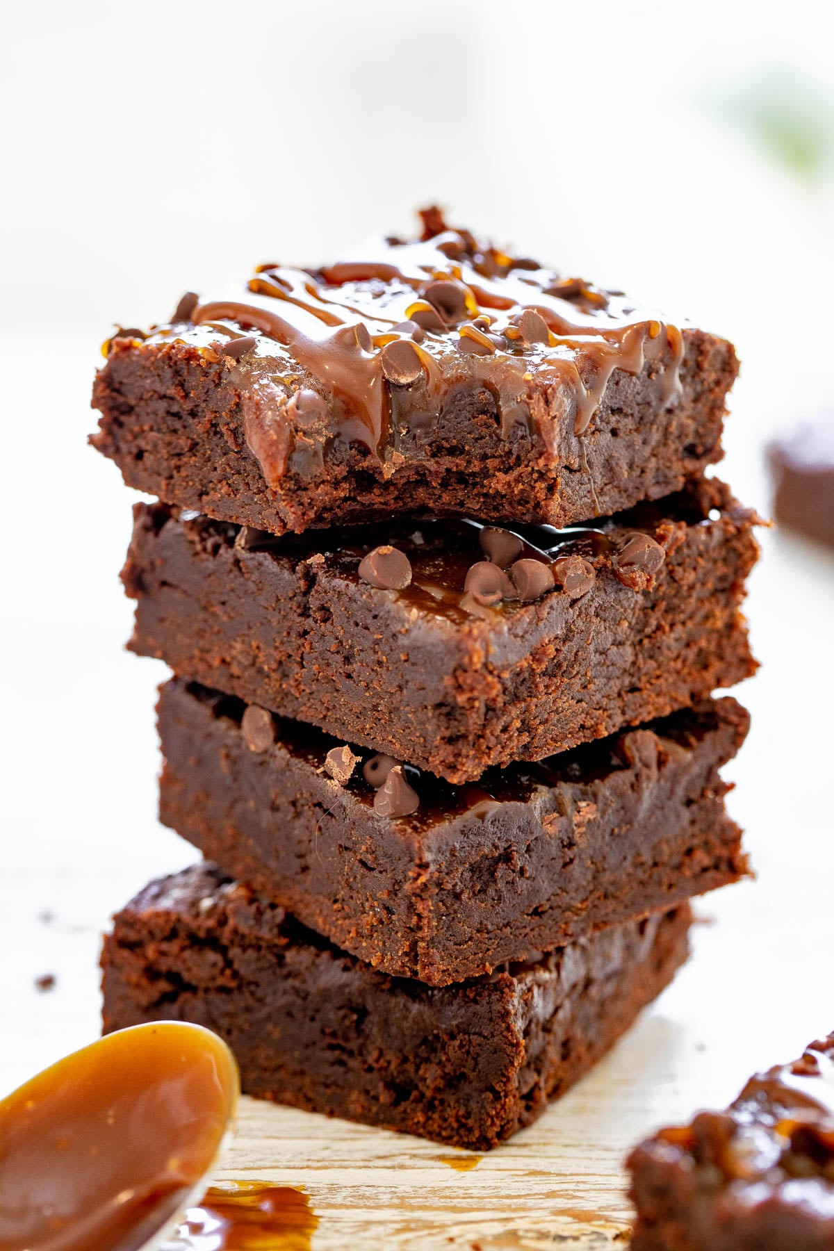 A stack of four chocolate brownies topped with caramel drizzle and chocolate chips, placed on a light-colored surface. A dollop of caramel is visible beside the brownies.