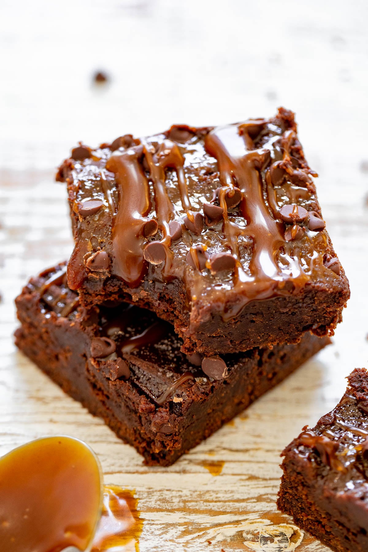 Two chocolate brownies stacked with caramel drizzled on top, set against a wooden surface. A spoon with caramel is nearby.