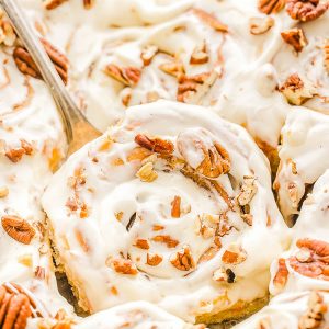 Close-up of frosted cinnamon rolls topped with chopped pecans, with a serving spoon lifting one.