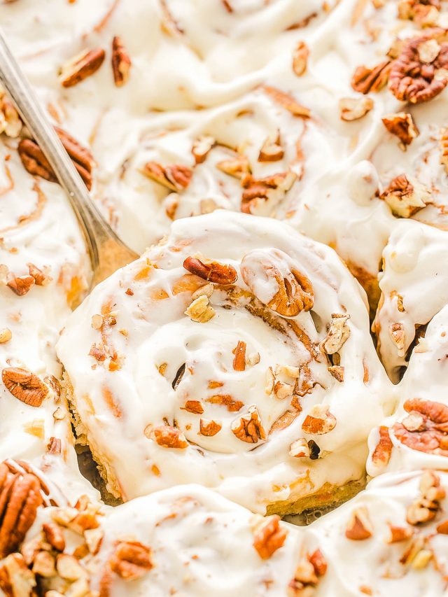 Close-up of frosted cinnamon rolls topped with chopped pecans, with a serving spoon lifting one.