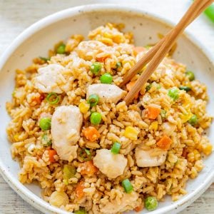 A bowl of chicken fried rice with peas, carrots, and corn, garnished with green onions. Chopsticks are placed on top of the dish.