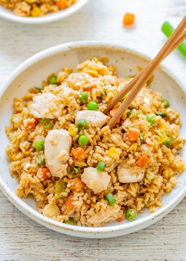 A bowl of chicken fried rice with peas, carrots, and corn, garnished with green onions. Chopsticks are placed on top of the dish.