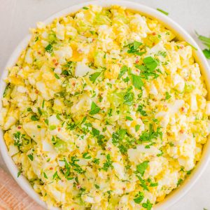 A bowl of egg salad garnished with chopped parsley, set on a light-colored surface.