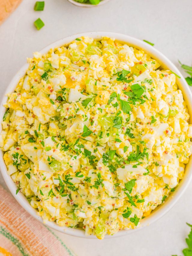 A bowl of egg salad garnished with chopped parsley, set on a light-colored surface.