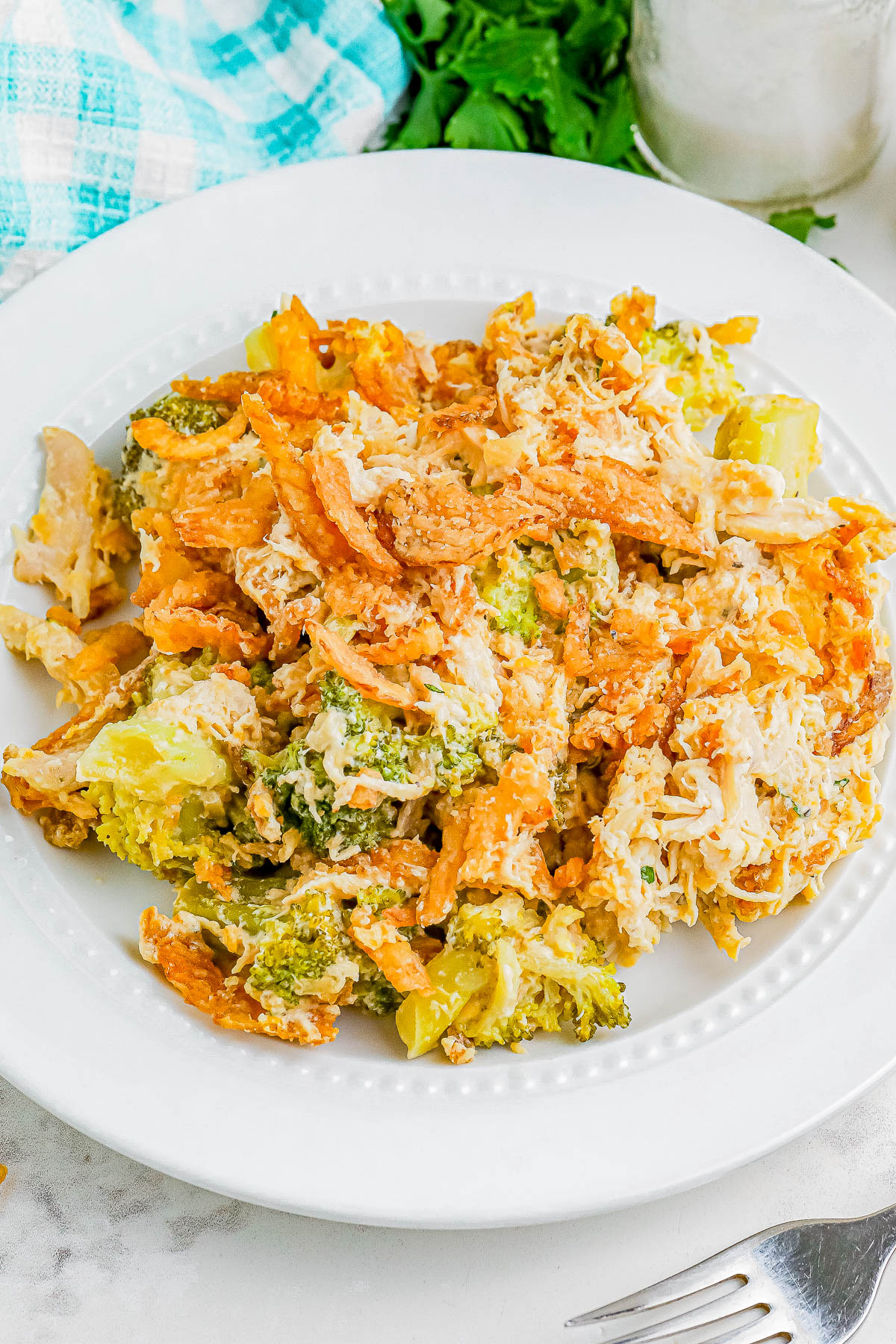 A white plate with a cheesy chicken and broccoli pasta bake, featuring shredded chicken, broccoli florets, and creamy sauce, on a marble surface with a fork nearby.