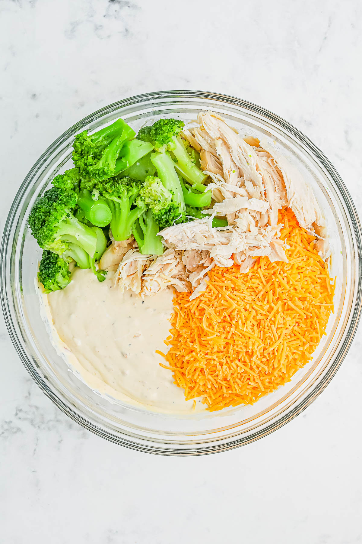 A glass bowl containing shredded chicken, chopped broccoli, grated cheese, and creamy sauce on a marble surface.